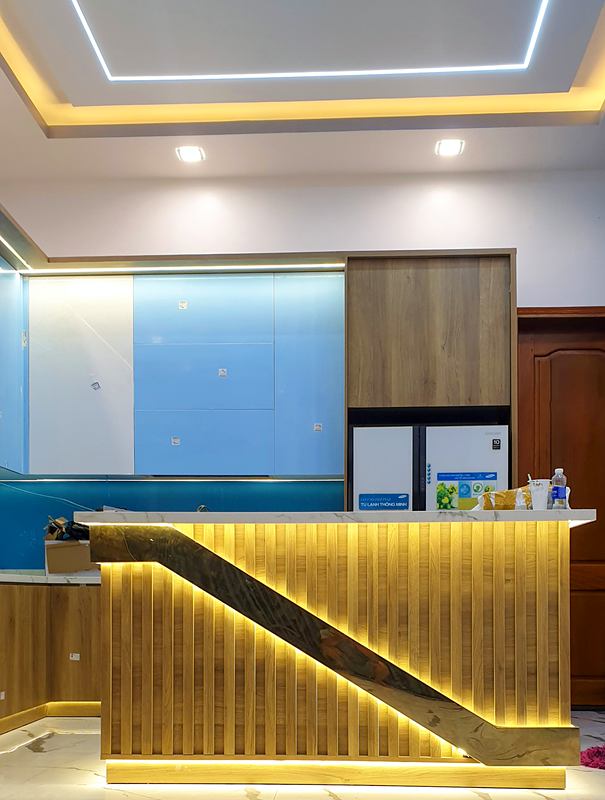 Quầy bar ốp lam gỗ có đèn hắt, phía sau là tủ bếp và kính ốp màu xanh